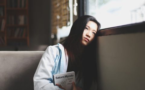 纪录片采访场景剧照，一位女性受访者坐在家中窗前接受采访，摄像灯打亮侧脸，背景是书柜